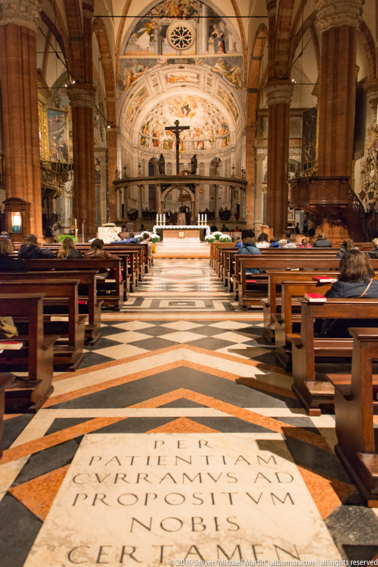 View down the cathedral nave over a geometric stone floor toward the altar, crucifix, and frescoed apse, with wooden pews and seated visitors.