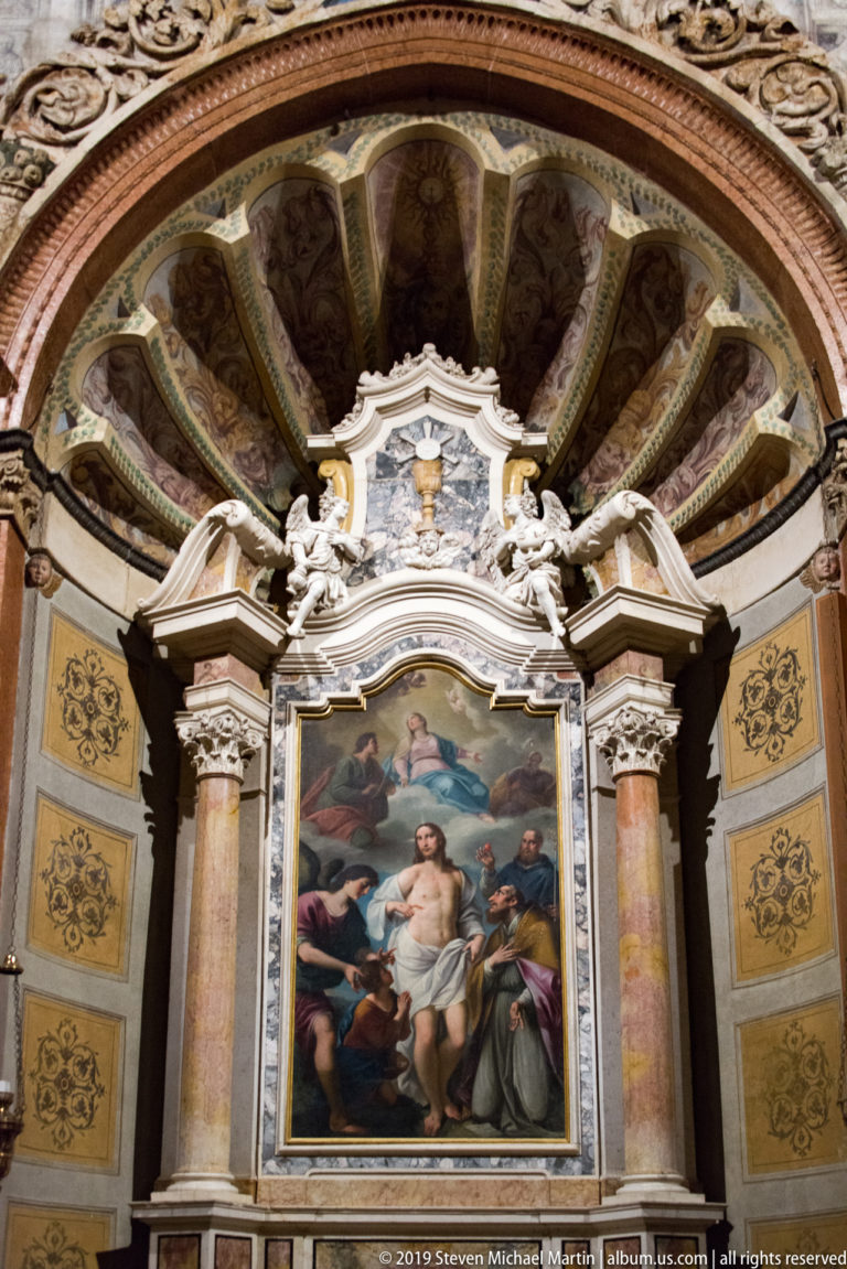 Religious altarpiece framed by columns and white sculpted ornament beneath a radiating, shell-like painted chapel vault.