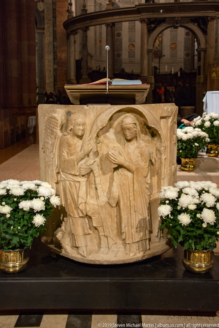 Close view of a pale stone altar relief carved with several draped figures, flanked by pots of white flowers.