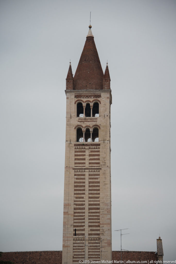 Upper San Zeno bell tower with paired arcades, striped masonry, corner pinnacles, and steep pyramidal roof.