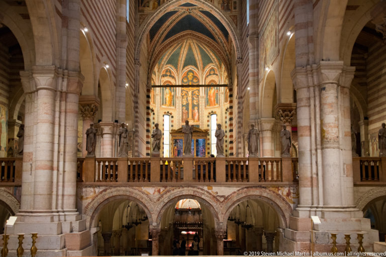 Frontal interior view of raised choir with statues, apse decoration, striped piers, and crypt arches below.
