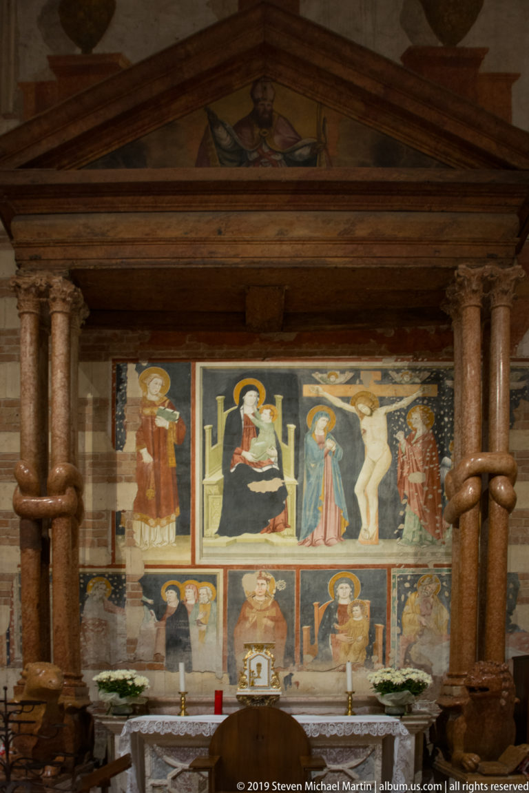 Small altar before colorful sacred fresco panels, framed by paired columns and a triangular pediment.