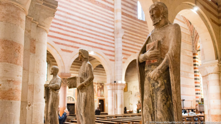 Standing stone religious figures beside striped Romanesque arches, with a bearded figure holding a rectangular object in front.