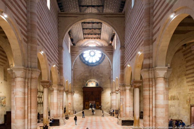 Long nave with striped piers and round arches leading toward a bright rose window above the entrance.