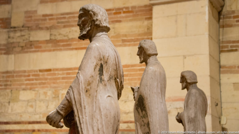 Profile view of several weathered stone religious statues receding through the striped nave interior.