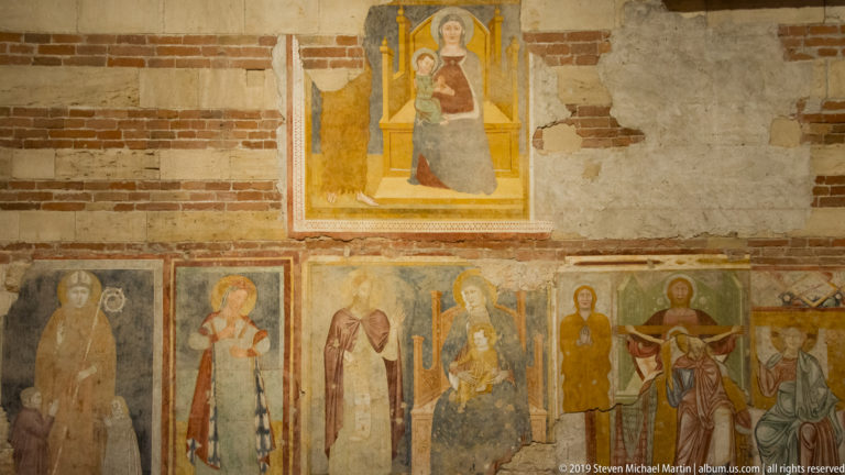 Seated Madonna and Child fresco above a row of haloed saints on a worn brick-and-plaster wall.