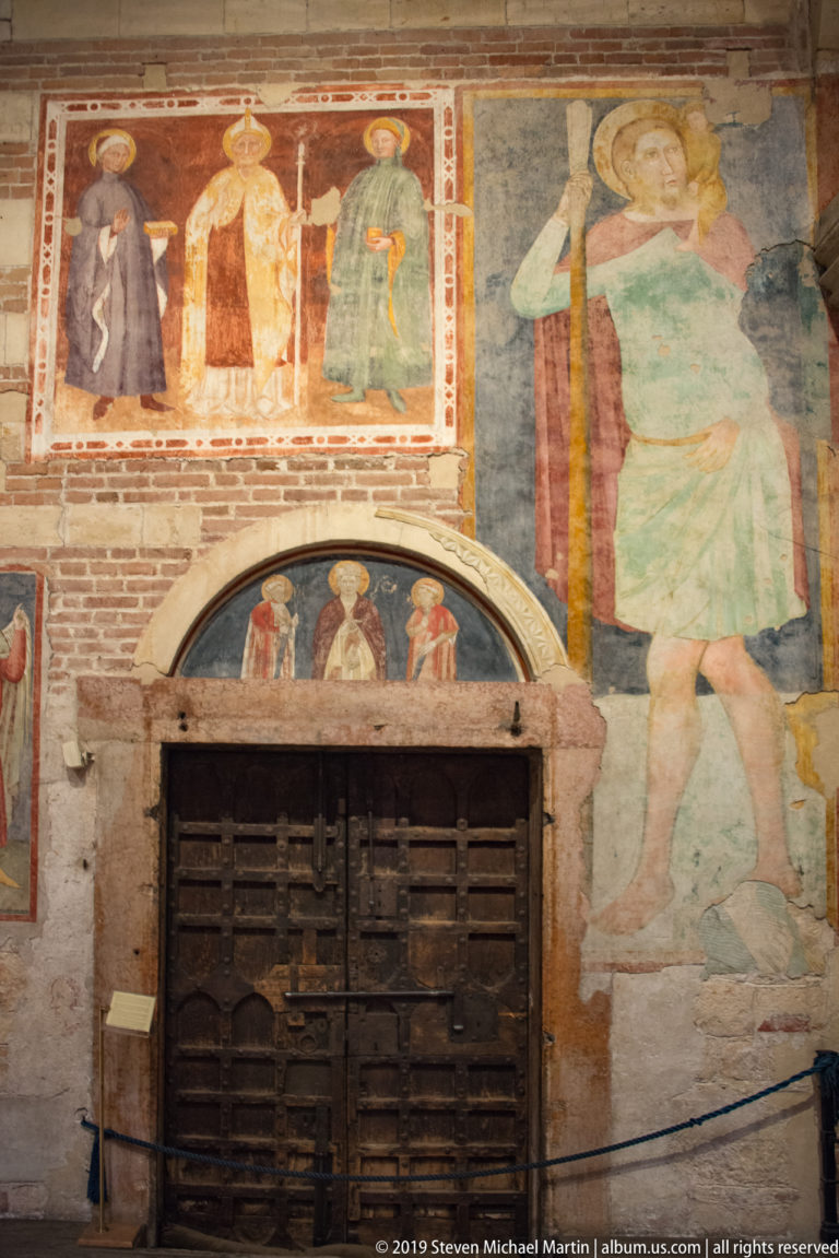 Large faded standing figure and smaller sacred fresco panels above a heavy wooden door on exposed brick.