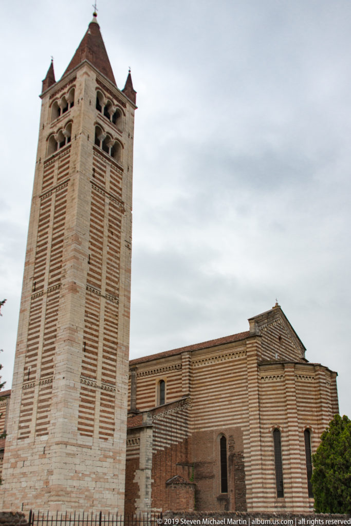 Tall striped campanile beside the lower brick-and-stone volumes of San Zeno under an overcast sky.