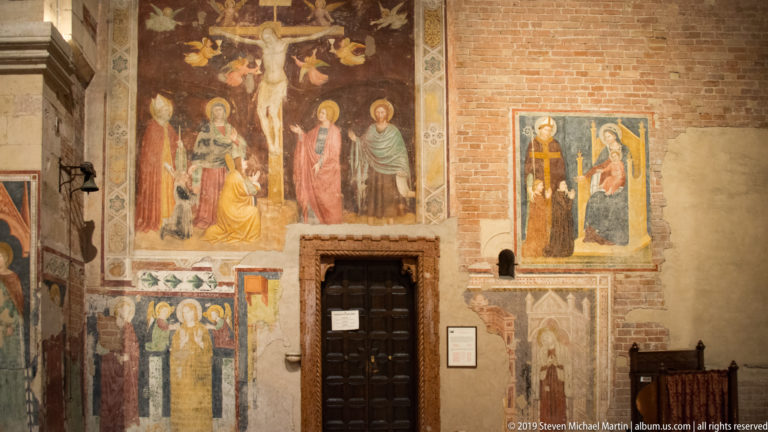 Fragmentary medieval frescoes, including a Crucifixion and saints, surrounding a dark wooden doorway.