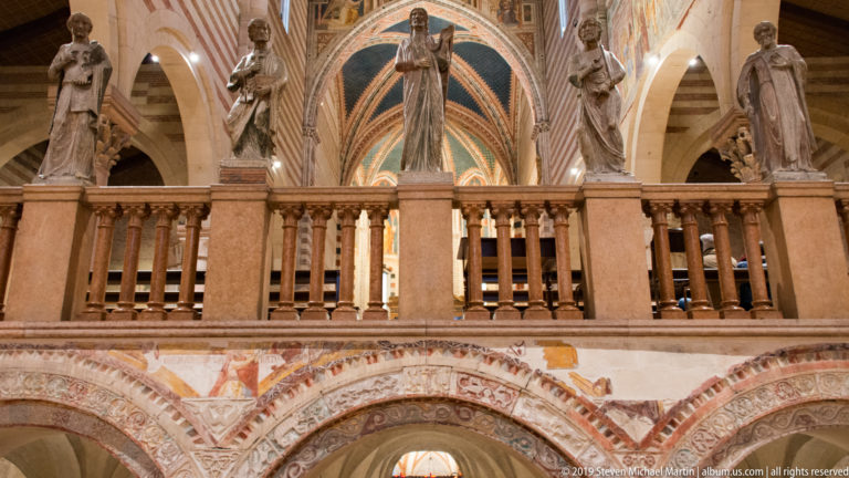 Stone religious statues line a balustrade above frescoed arches, with the painted apse vault behind.