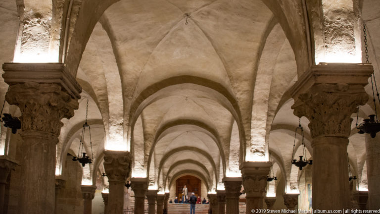 Long low crypt interior with rows of stone columns, groin vaults, hanging lamps, and distant visitors.