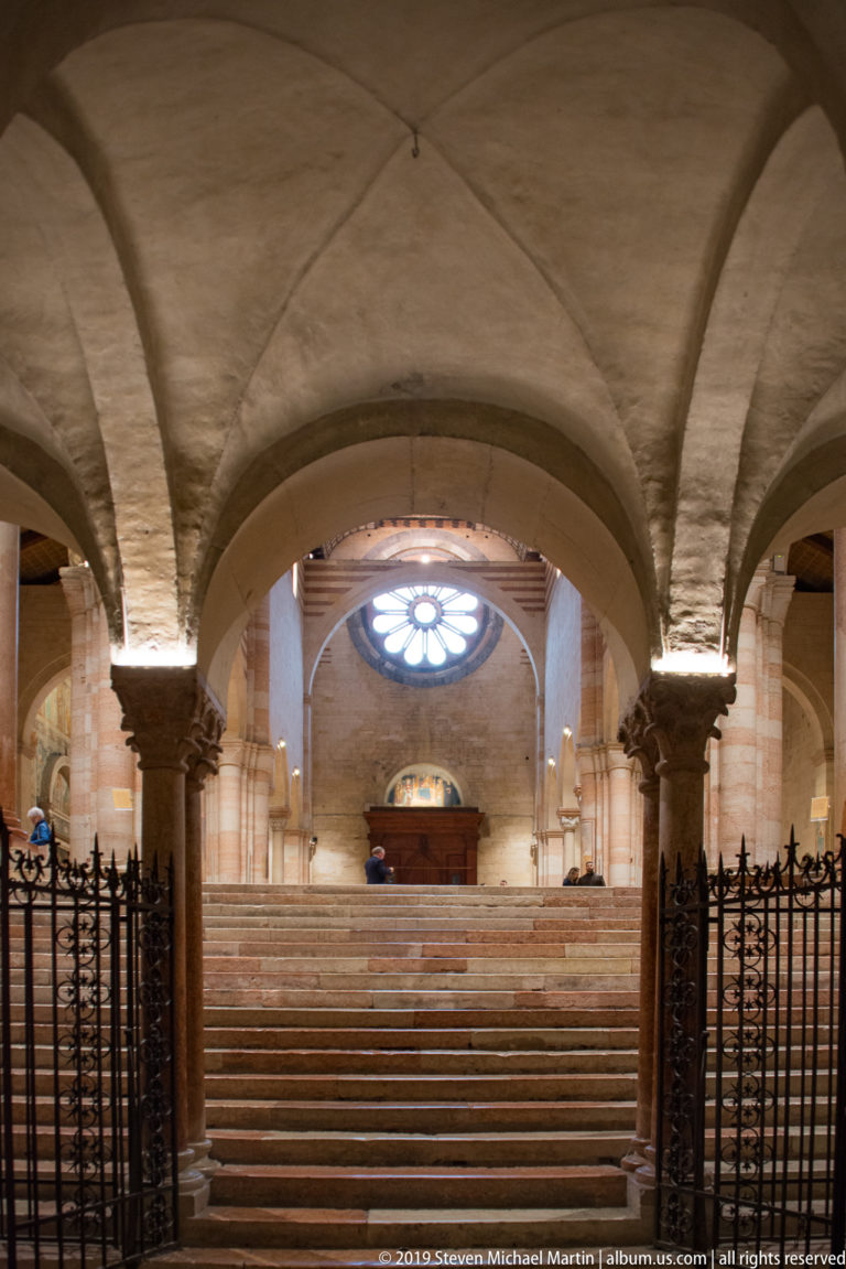 Symmetrical crypt view with open iron gates, stone staircase, columns, and circular window beyond.
