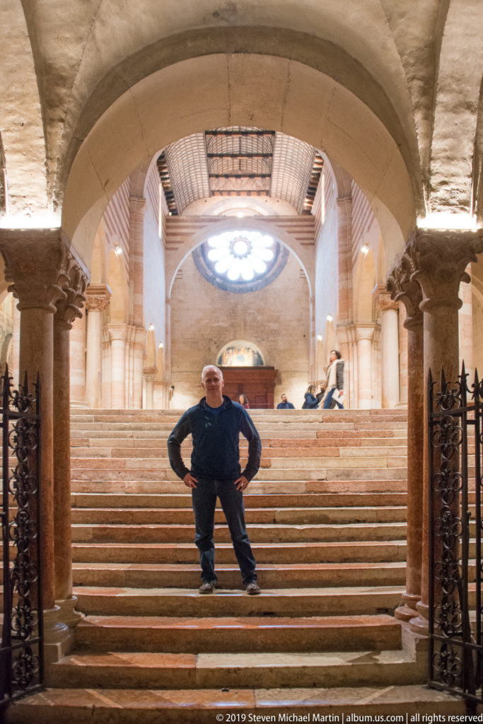 Visitor standing on stone steps between crypt columns, with iron gates and a bright circular window above.