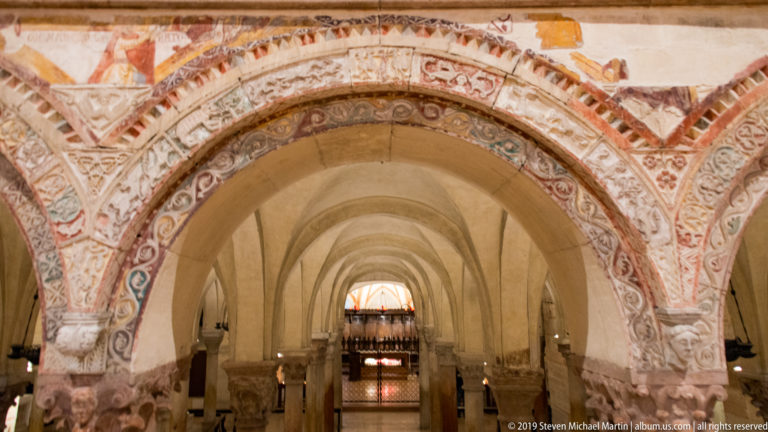 Frescoed arch opening into a low vaulted crypt with repeated arches receding into the distance.