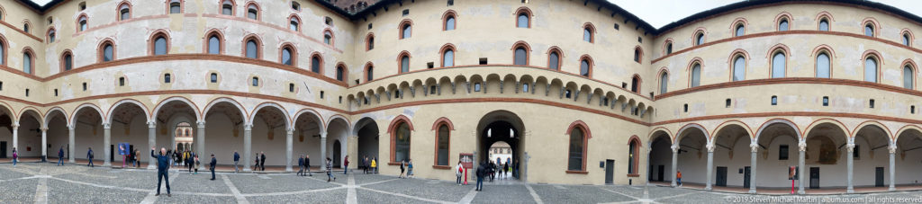 Wide view of San Zeno cloister with round-arched arcades, pale walls, red-brick bands, and visitors.