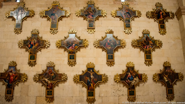 Multiple ornate painted crosses arranged in rows on a pale stone wall inside the basilica.
