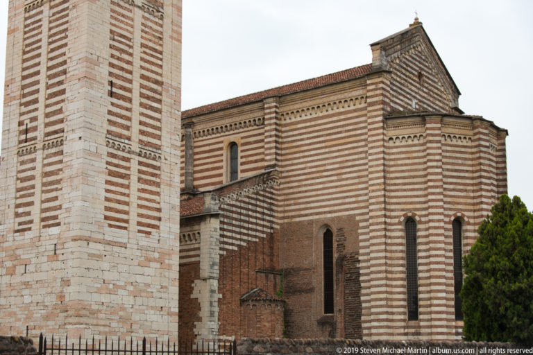 Close exterior view of San Zeno with alternating brick-and-stone bands, narrow windows, and stepped rooflines.