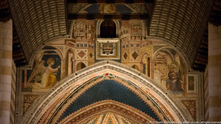 Frescoed upper wall above a pointed sanctuary arch, with large figures, geometric patterns, and heraldic motifs.