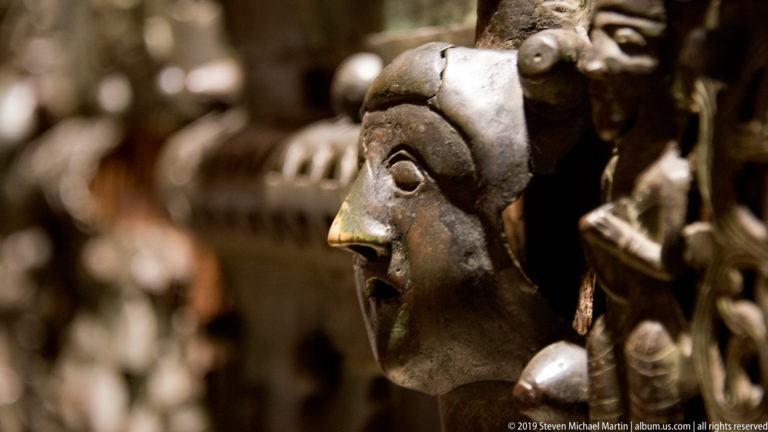Close-up of a dark cast bronze head projecting from the door, with blurred relief figures behind it.