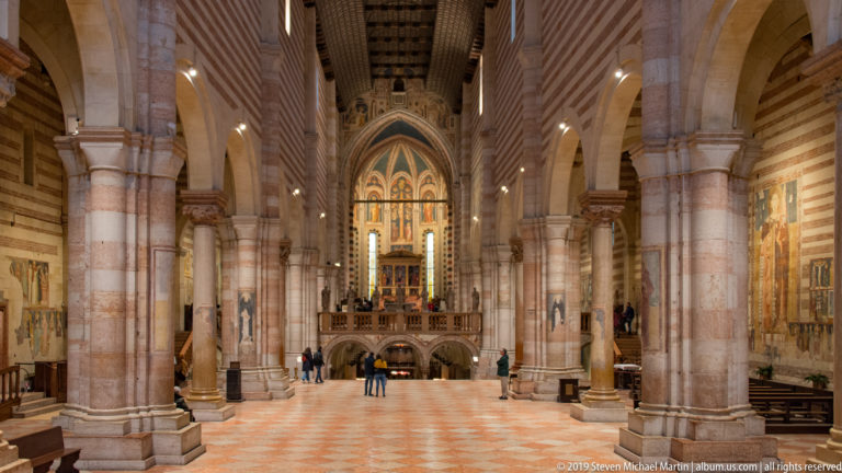 Long central nave with striped piers, patterned floor, timber roof, and bright raised sanctuary at the far end.