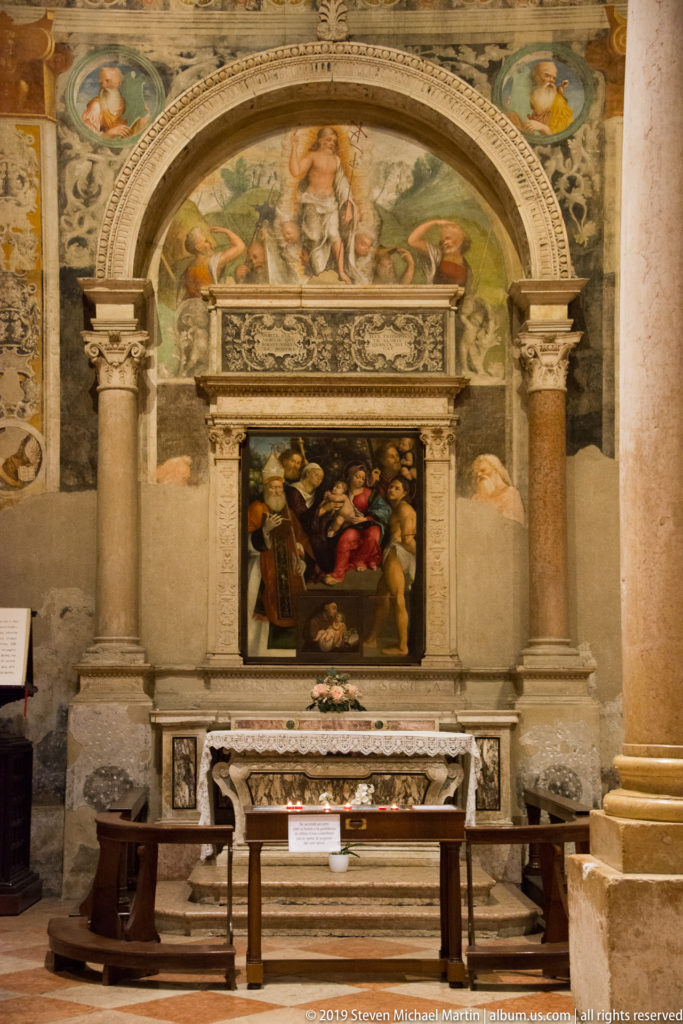 Side altar with a framed religious painting set within a frescoed arched niche and flanked by columns.
