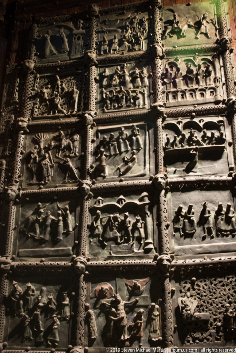 Section of bronze church doors with many relief panels, ornamental bands, and varied dark and greenish patina.