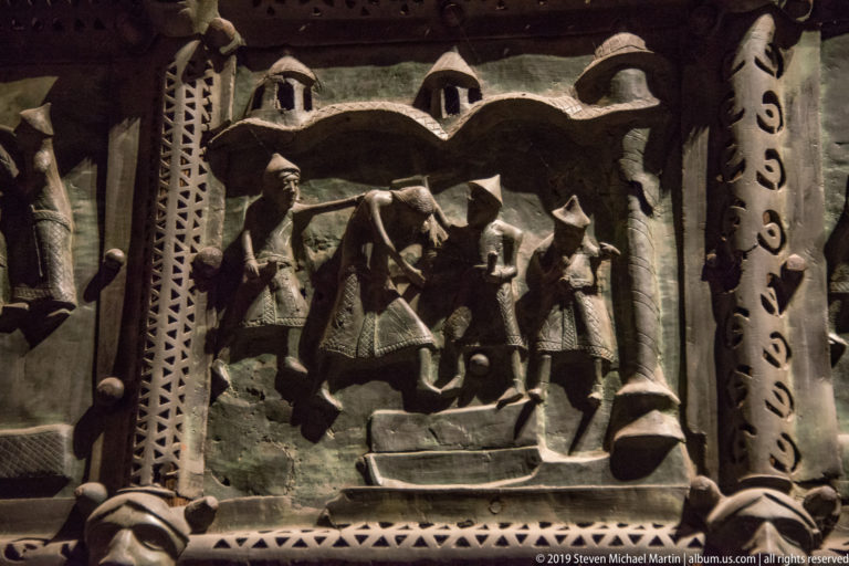 Bronze relief panel with figures beneath a stylized architectural canopy, bordered by patterned metal strips.