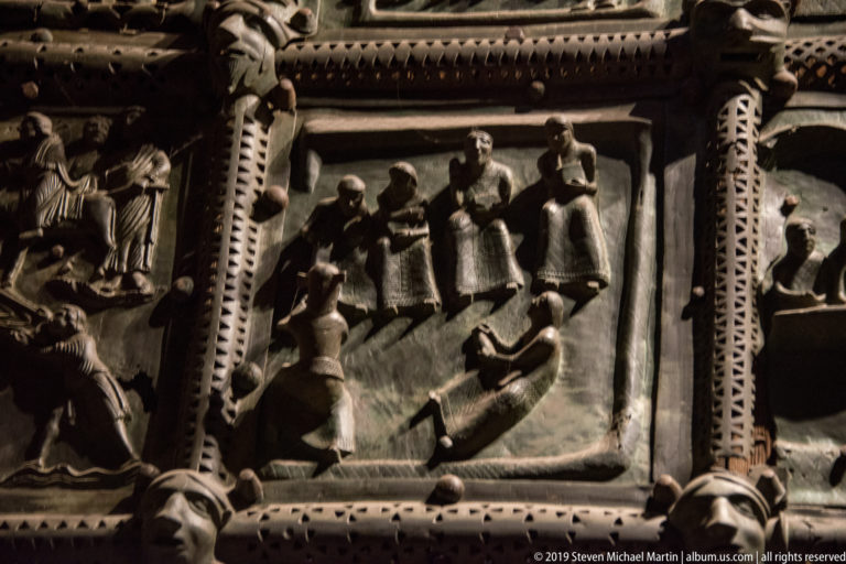Single bronze door relief with several standing figures and another figure below, surrounded by patterned metal bands.