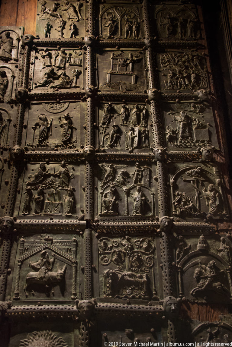Oblique close-up of bronze church-door panels filled with small narrative relief scenes and patterned borders.