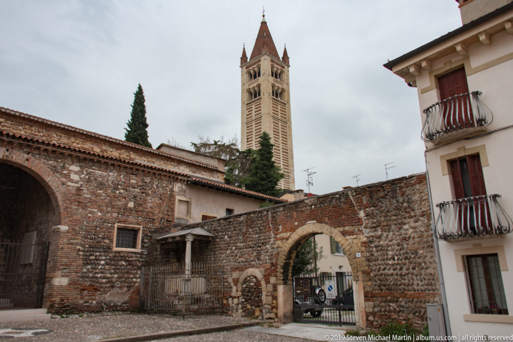 San Zeno bell tower above old brick-and-stone walls and an arched gateway under a cloudy sky.