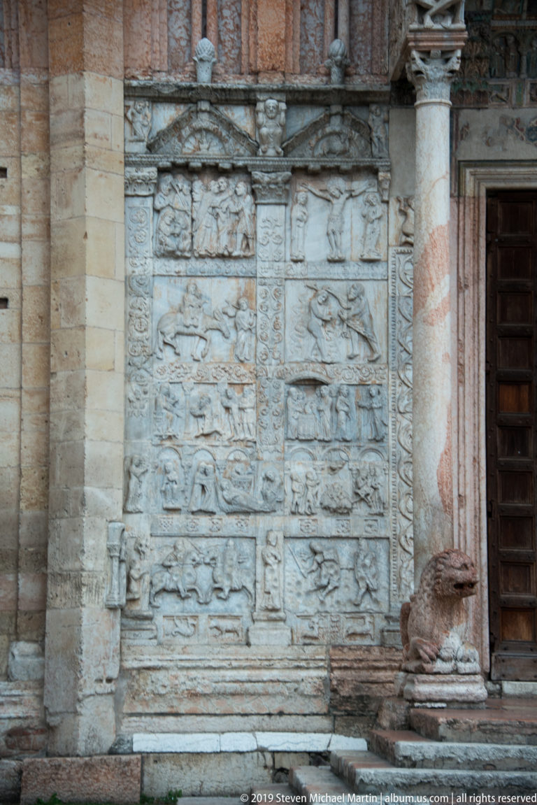 Lower carved stone relief panels beside the portal, next to a spiral-banded column and wooden door.