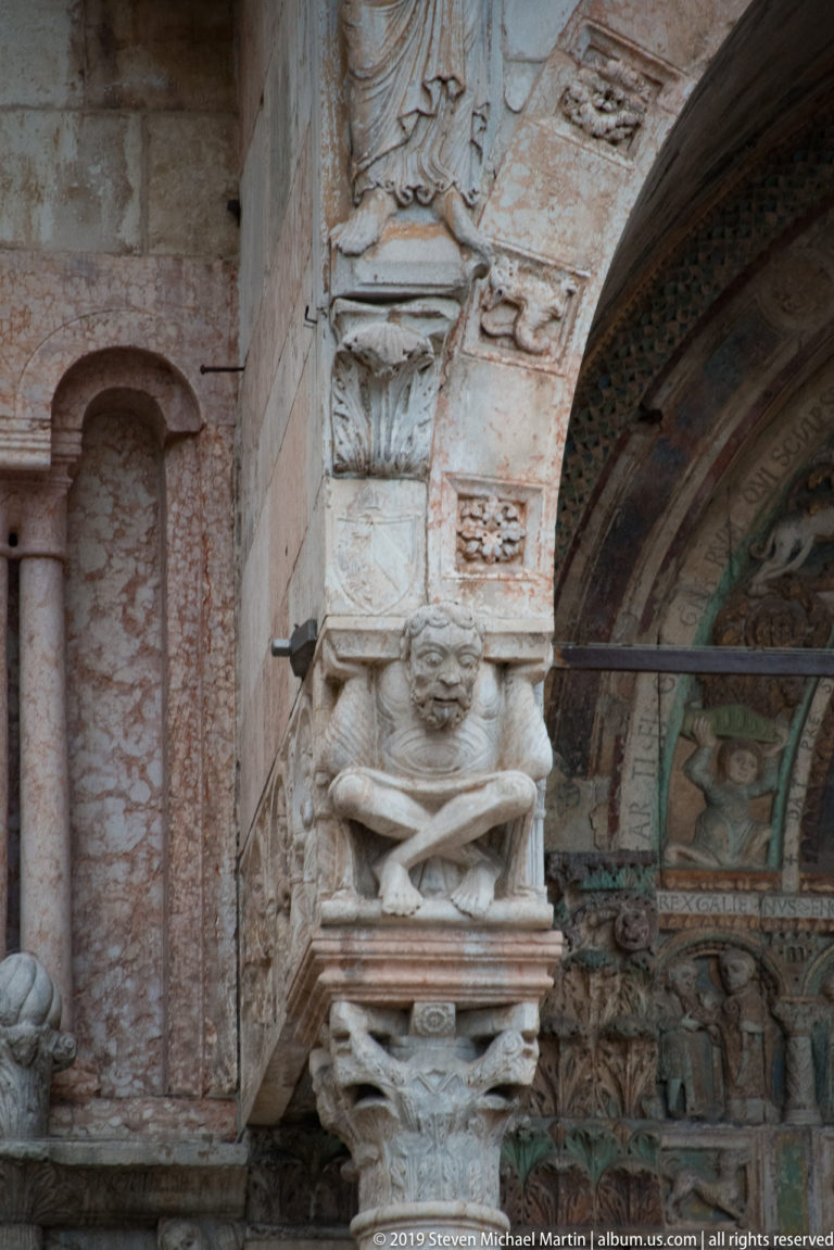 Close-up of a figurative carved stone capital at the portal with part of the painted tympanum behind it.