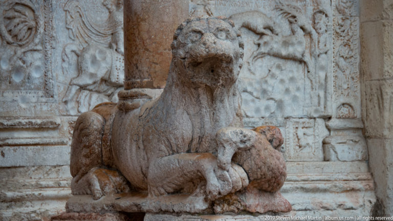 Weathered stone lion at the portal, crouching on a plinth beside carved façade reliefs.