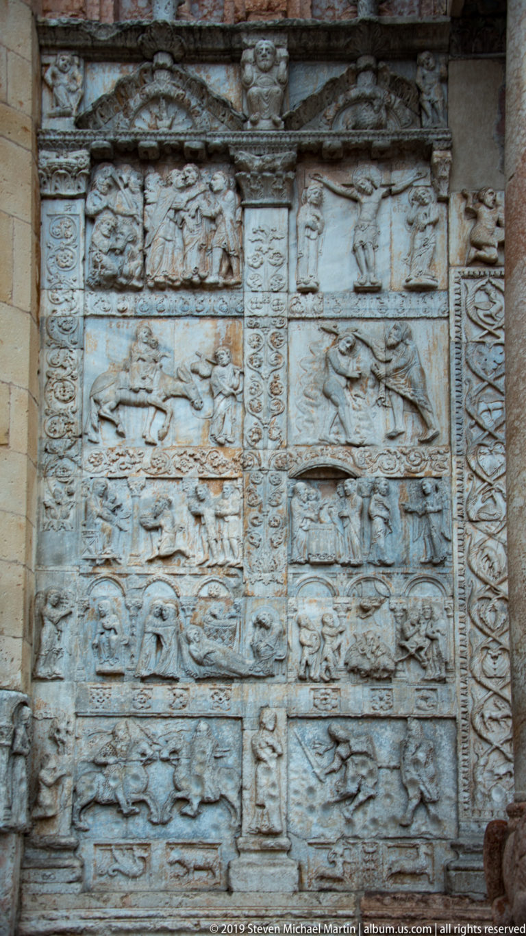 Weathered stone relief panels beside the church portal, arranged in stacked narrative scenes with decorative borders.