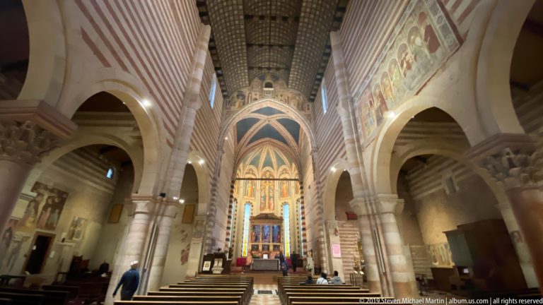Centered nave view toward the bright raised sanctuary, framed by striped piers, arches, wall paintings, and a dark timber ceiling.