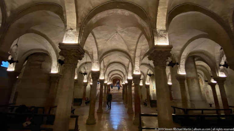 Low crypt interior with many stone columns, rounded arches, ribbed vaults, and a central stair in the distance.