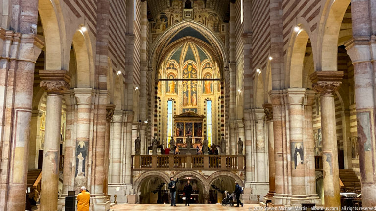 Long basilica nave with striped stone-and-brick piers leading to a raised, painted presbytery above lower arches.