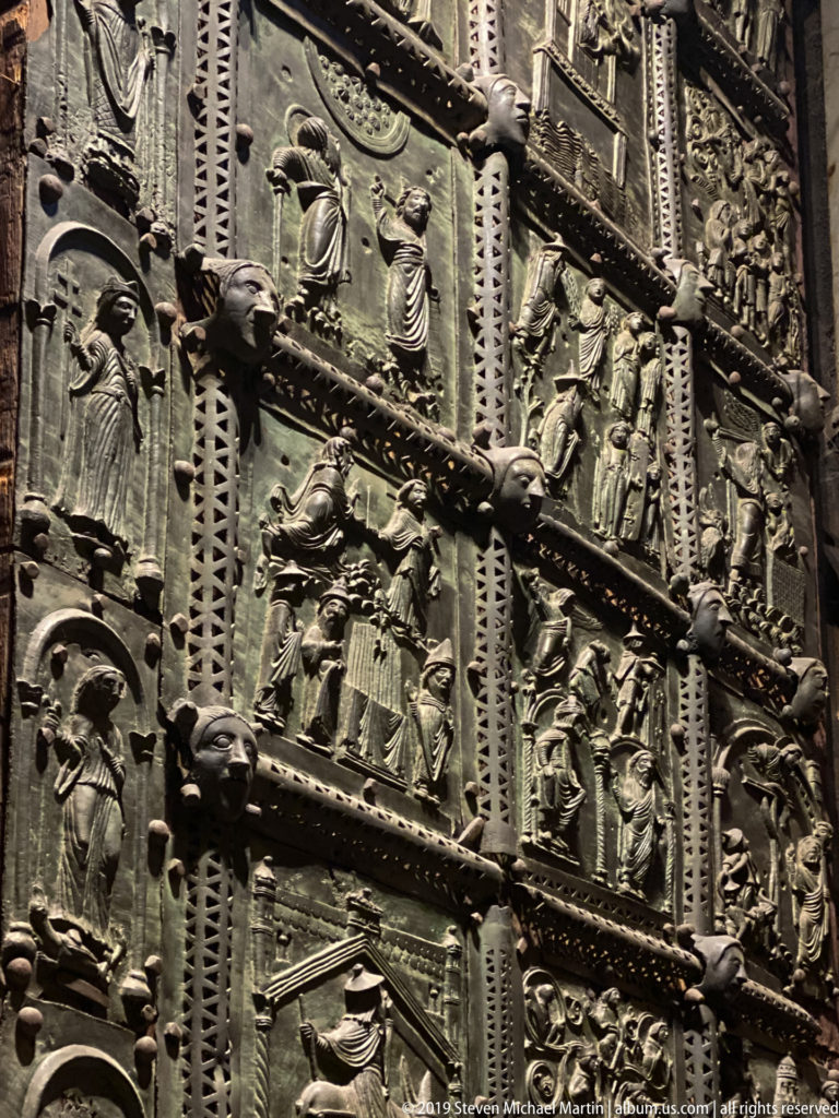 Close-up of dark bronze church doors divided into narrative relief panels with patterned metal borders.