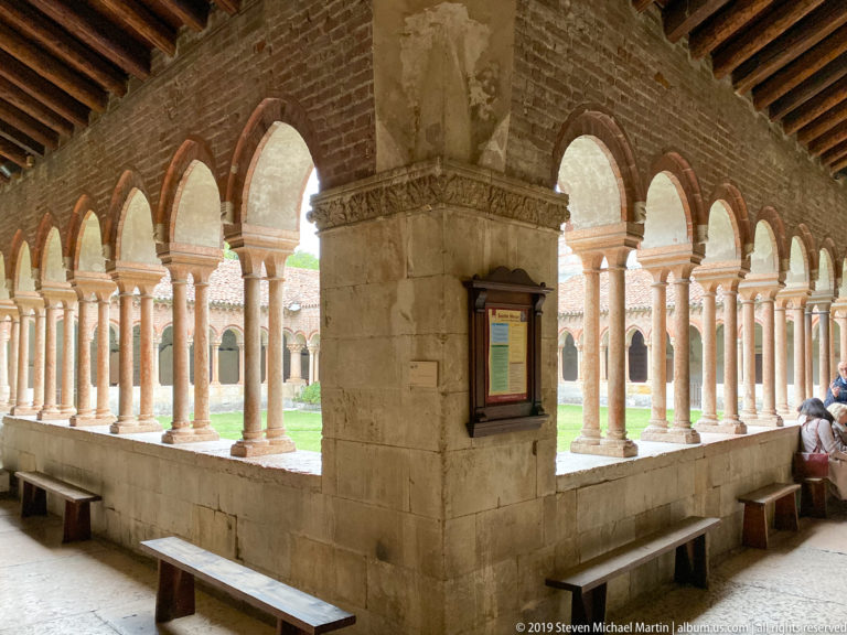 Covered cloister walk with rows of small arches and columns opening onto a grassy courtyard.