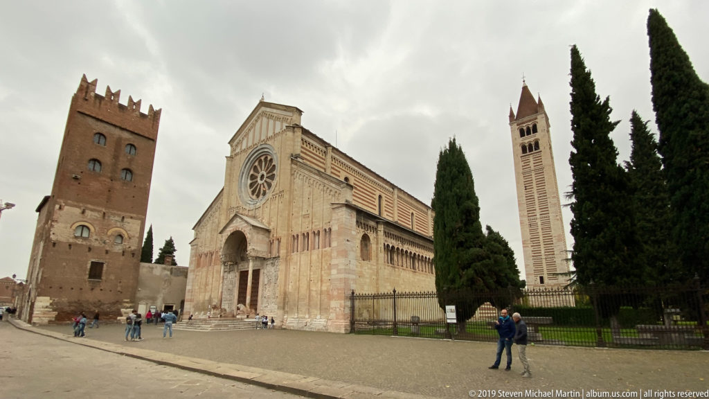 Wide view of San Zeno’s façade between a crenellated brick tower and a tall striped bell tower.