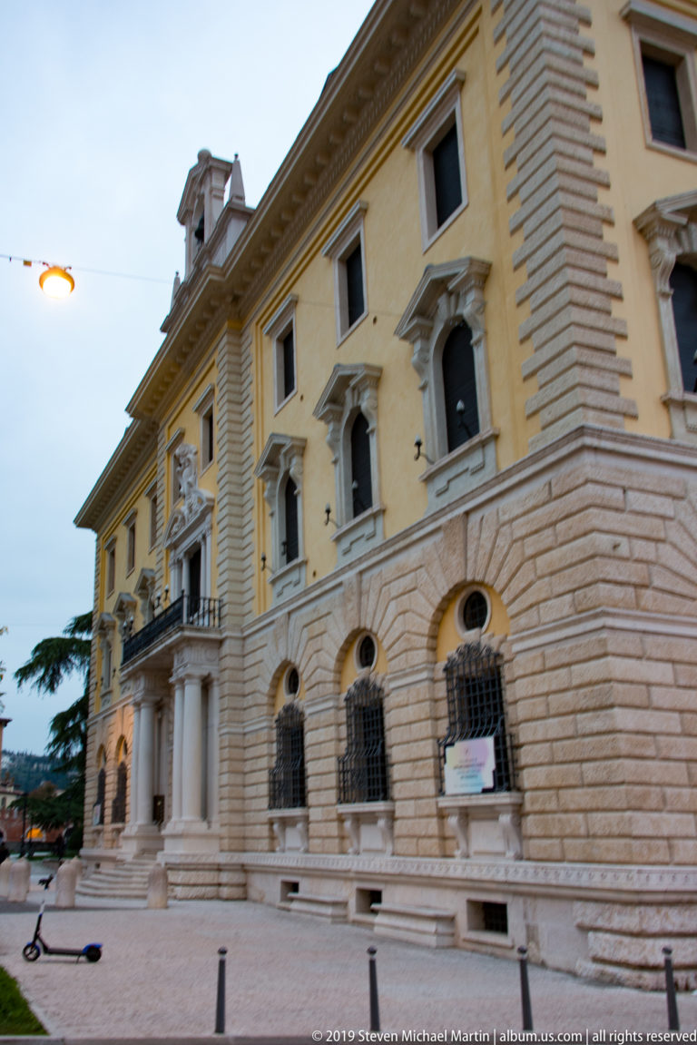 Angled dusk view of a monumental pale civic building with rusticated base, pedimented windows, pilasters, and projecting columned entrance.
