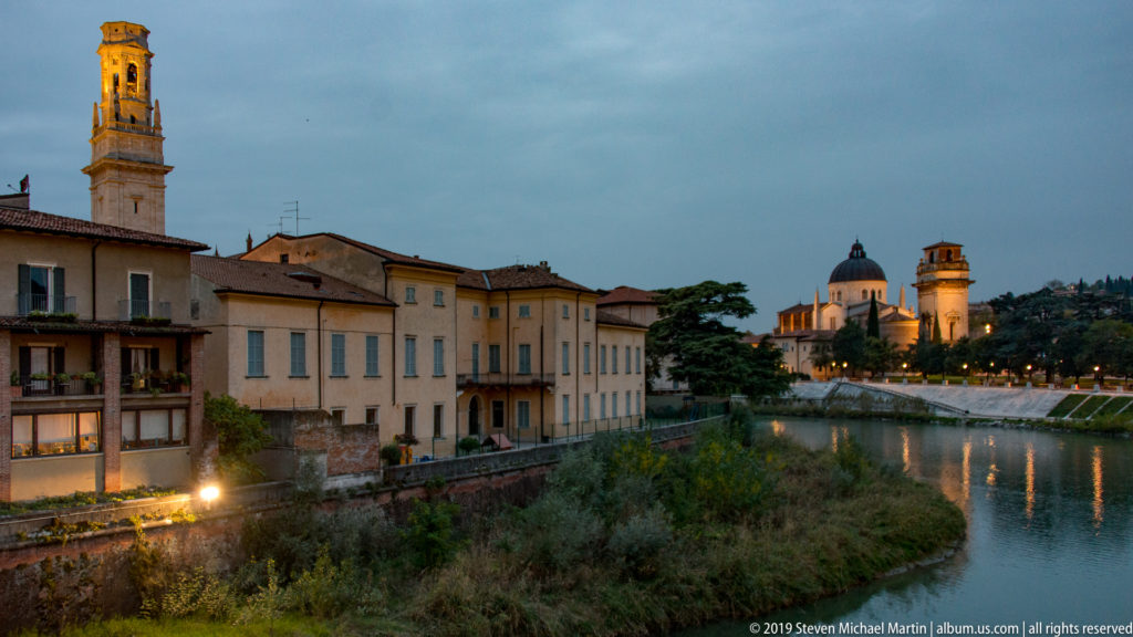 Adige river bend with vegetated bank, riverside houses, and illuminated San Giorgio in Braida dome and bell tower in the distance.