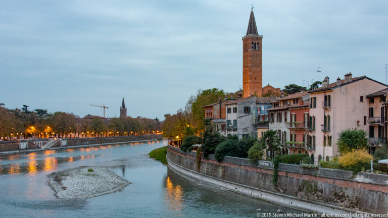 Broad Adige river bend with exposed gravel bar, riverside houses, tall brick campanile, trees, and a line of evening lights.