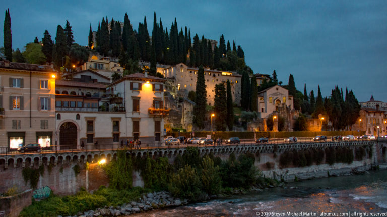 Dusk view along the Adige toward the cypress-covered Castel San Pietro hillside, riverside buildings, and glowing streetlights.