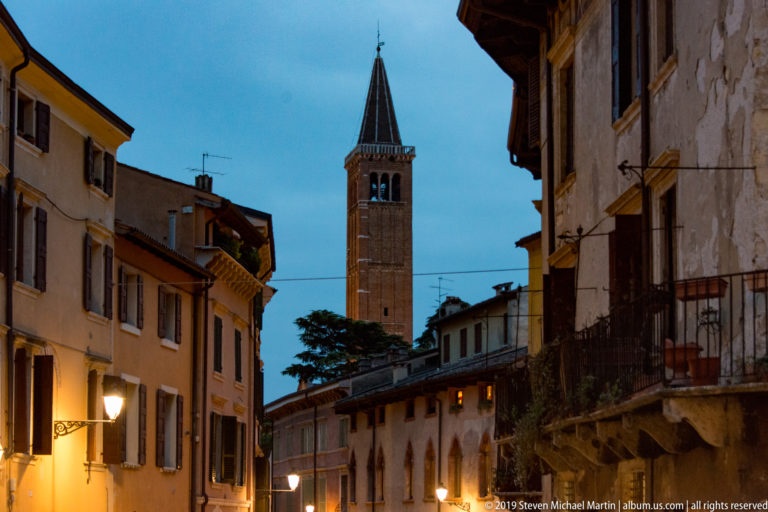 Tall brick bell tower with pointed roof above a narrow Verona street lined with stucco houses, balconies, and warm lamps.