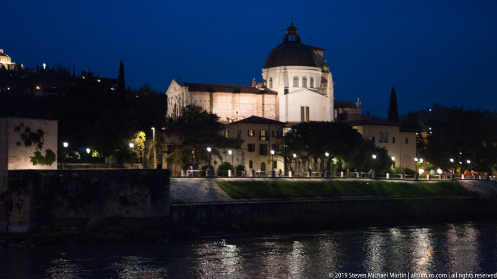 Closer night view of San Giorgio in Braida’s bright drum and dark dome above the Adige, trees, and reflected river lights.