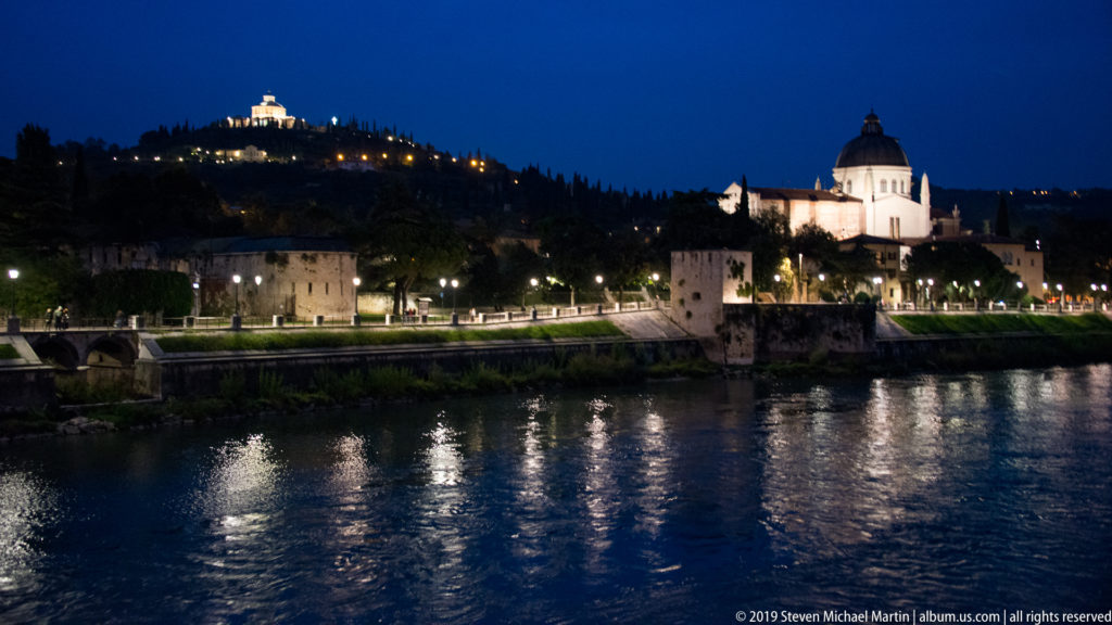 San Giorgio in Braida illuminated across the Adige, with domed church, riverbank lights, hillside, and reflections on the water.