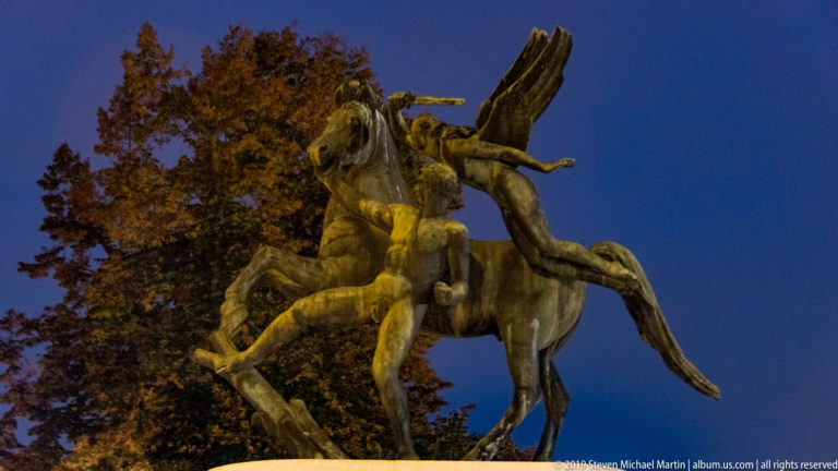 Low-angle view of a bronze equestrian sculpture with rearing horses and elongated riders against blue sky and autumn trees.