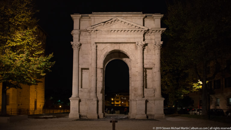 Illuminated Arco dei Gavi with single arched opening, classical columns, capitals, and pediment against a dark night setting.
