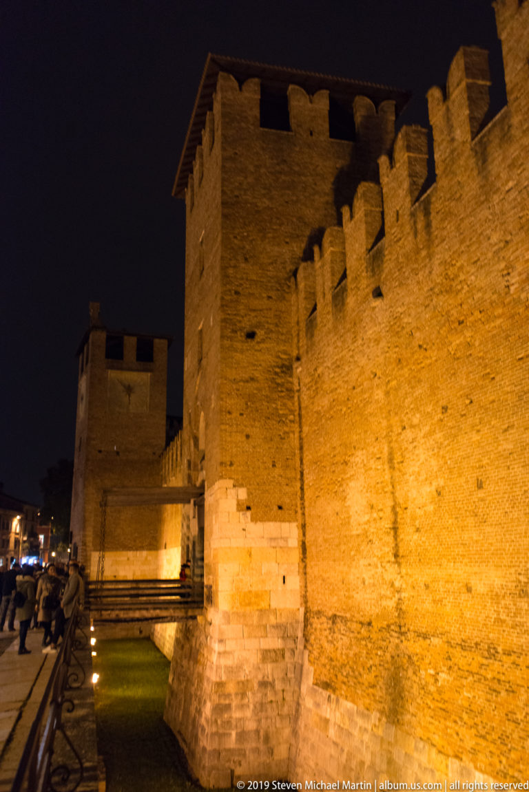 Oblique night view along Castelvecchio’s floodlit brick outer walls, square towers, crenellations, and pale stone base.